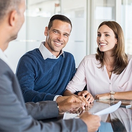 A man and woman, both smiling, sit across a table from another person in an office setting. They appear engaged in a friendly conversation about computer repair, with the man in a blue sweater and the woman in a light shirt. Papers are on the table. computer repair