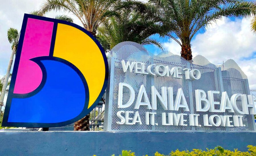 A vibrant, colorful sign with a large stylized D and the text "Welcome to Dania Beach. Sea it. Live it. Love it." set against a backdrop of palm trees and a partly cloudy sky reminds visitors that even paradise sometimes needs computer repair support. computer repair
