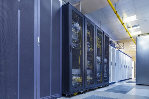 A row of server racks in a data center, housing various electronic equipment crucial for computer repair. The room has a tiled floor and a ceiling with piping and fluorescent lights. Cables and connections are visible inside the transparent doors of the racks. computer repair