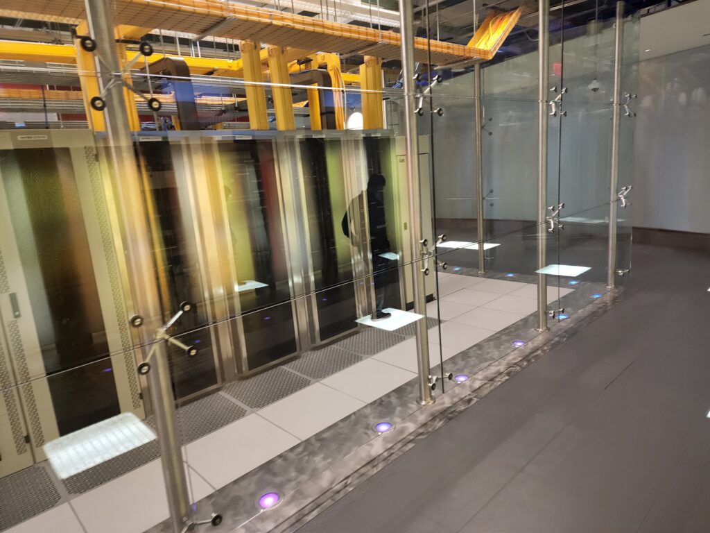 A modern data center with rows of server racks enclosed in glass hums with activity. Bright lighting illuminates the floor, where a skilled technician is faintly visible, adding to the high-tech and secure atmosphere. Yellow structural elements above hint at computer repair capabilities within. computer repair
