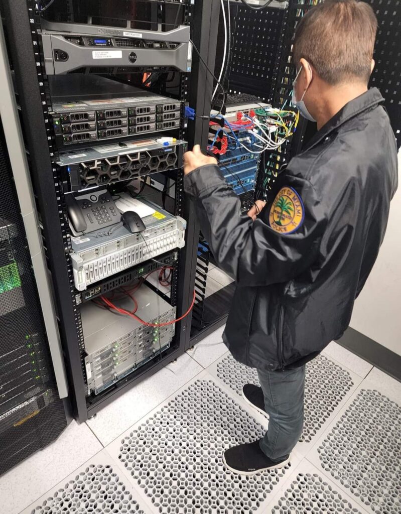 A person in a black jacket is working in a server room, skillfully adjusting cables on a rack of computer servers as part of a computer repair. The floor is covered with vented tiles, and various wires are visible connecting the equipment. computer repair