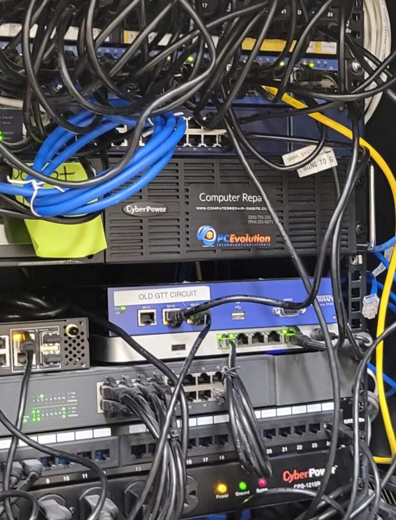 A photo of a cluttered network server rack with tangled cables, reminiscent of a computer repair project. Various pieces of networking equipment, including switches and power units, are visible. Labels such as CyberPower and OLD GTT CIRCUIT can be seen on some devices. computer repair