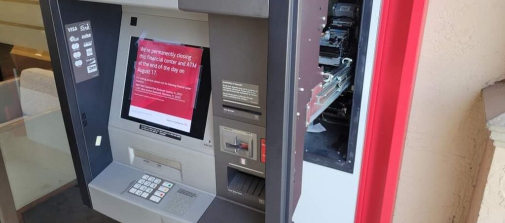 An ATM with its screen displaying a closing message stands with the right side panel open, revealing internal components as if awaiting computer repair. A nearby window reflects the warm interior light. computer repair