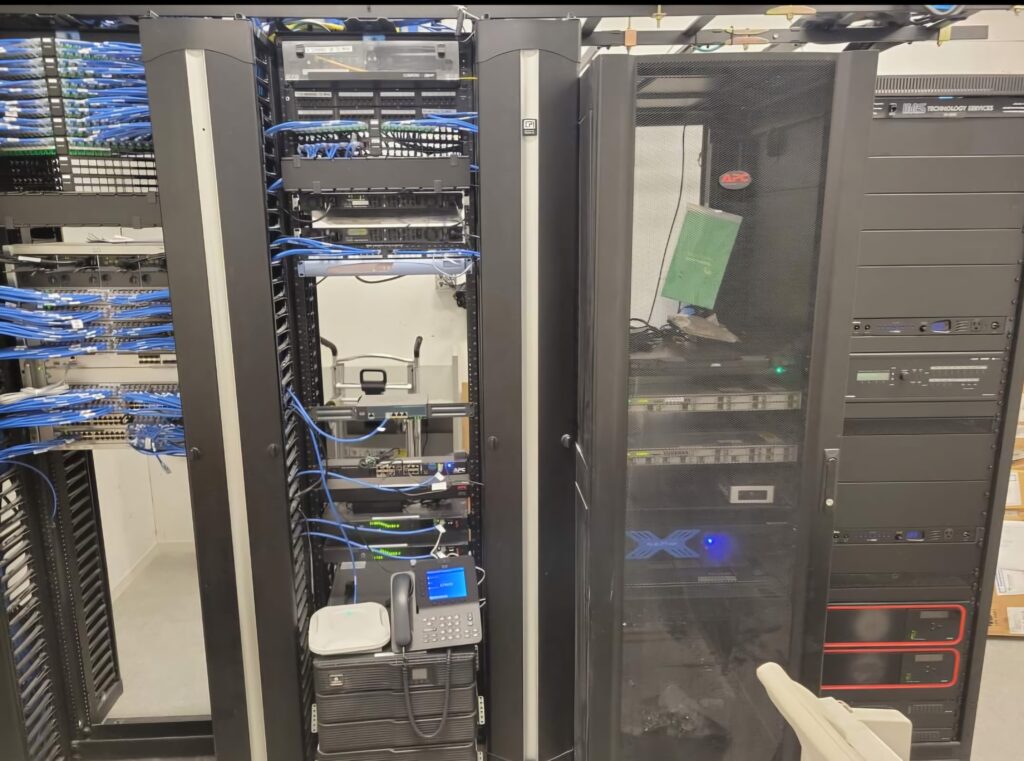 A server room with several racks filled with network switches, servers, and a variety of cables awaits a computer repair team. A phone and some equipment are on a cart in front of the racks. The room is well-organized with blue and black cables neatly arranged. computer repair