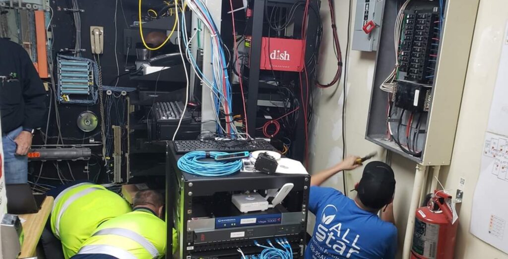 Two workers are setting up network servers and equipment in a cluttered room filled with wires and computer hardware, resembling a computer repair hub. One is wearing a green safety vest, and the other is in a blue shirt. A fire extinguisher is visible on the right. computer repair