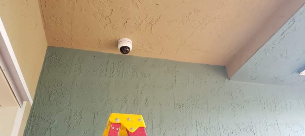 A security camera is mounted on the ceiling in a corner, with beige and green textured walls, quietly observing. Below it stands a red and yellow step ladder, reminiscent of a diligent tool awaiting its next task, much like one in a computer repair shop. computer repair