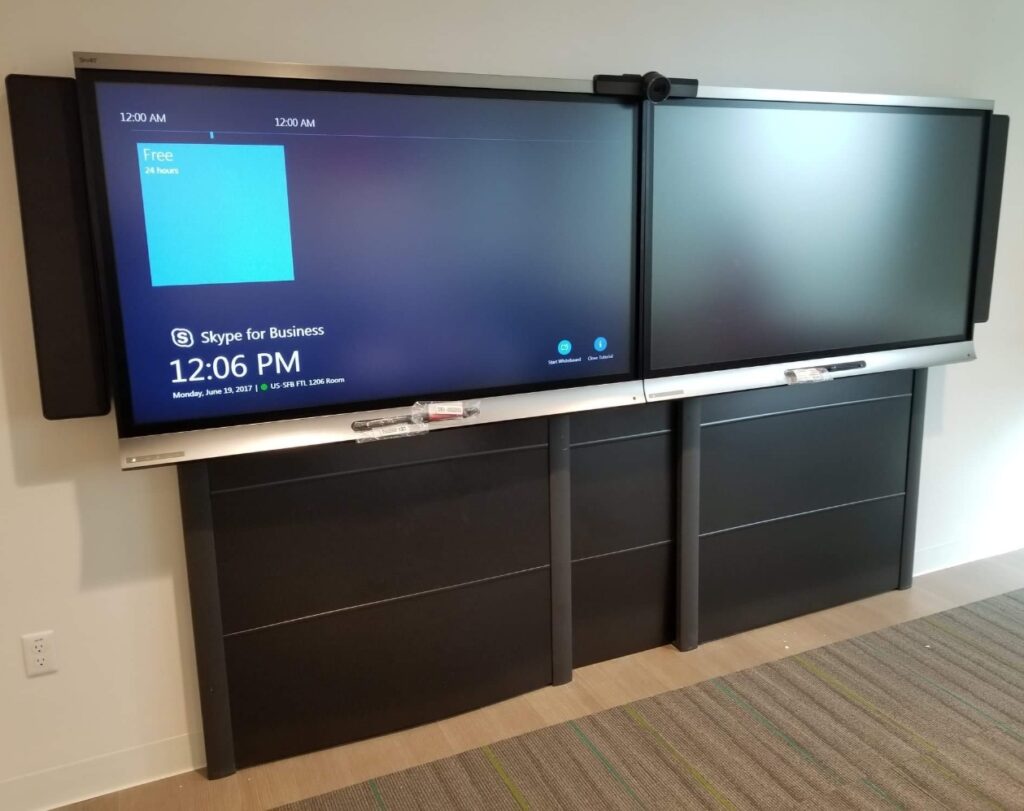 A conference room with a dual-screen setup displaying Skype for Business, where a technician often performs computer repair. The left screen shows a meeting schedule and the time as 12:06 PM. The room is carpeted with a stripe pattern, and the screens are mounted on a black cabinet. computer repair