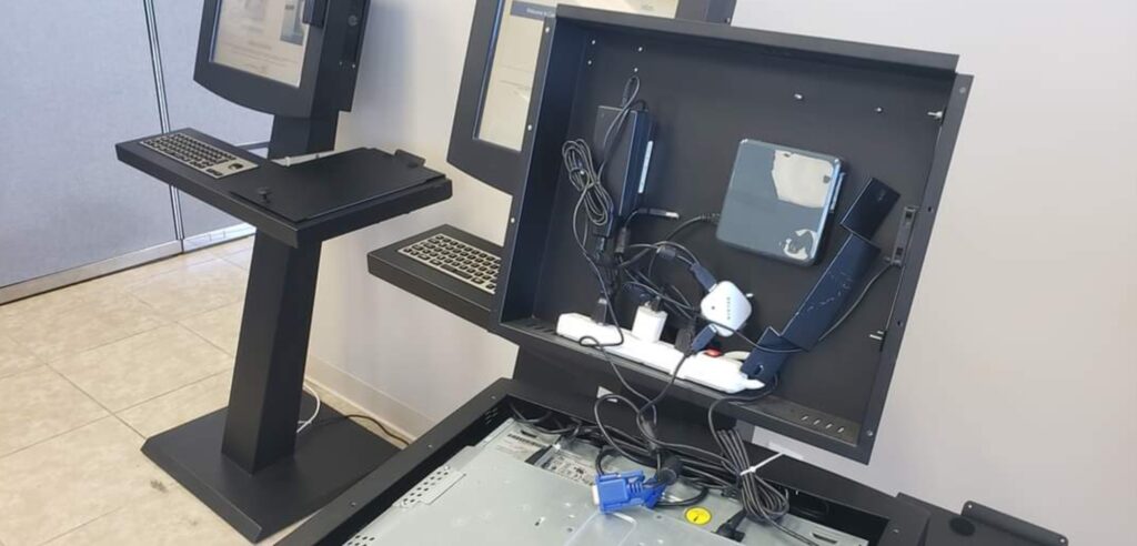 An open electronic kiosk with exposed wires and components, displaying its internal hardware. Two other kiosks stand nearby with keyboards attached. The floor is tiled and a partial view of a partitioned wall is visible in the background.