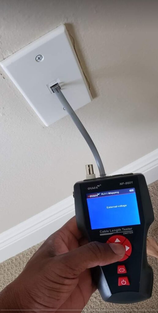 A hand is holding a cable length tester with a blue screen displaying External voltage. The tester is connected to a wall Ethernet port via a gray cable. The wall is light-colored, and the floor is carpeted.