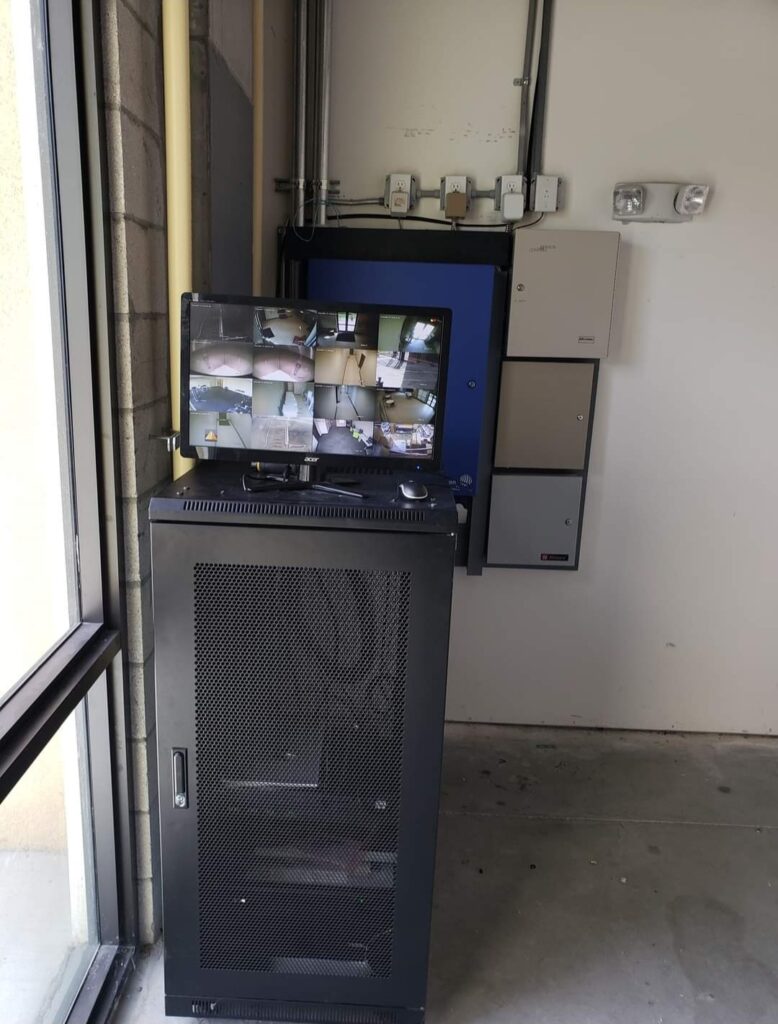 A security system with a black metal cabinet and a monitor displaying multiple camera feeds is set up in a room with concrete walls and a window. Electrical panels are mounted on the wall nearby.