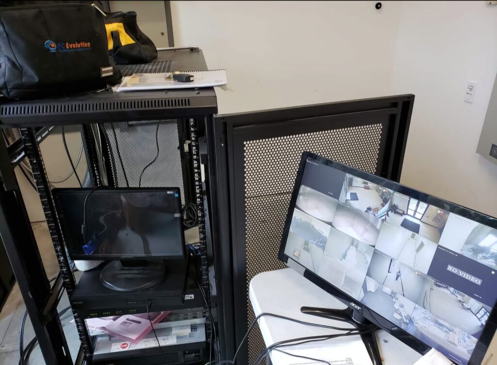 A room with a computer setup, including several monitors displaying multiple security camera feeds. Nearby, a black equipment rack holds various technical gear, and a black bag is placed on top. Papers and a notebook are on the table.