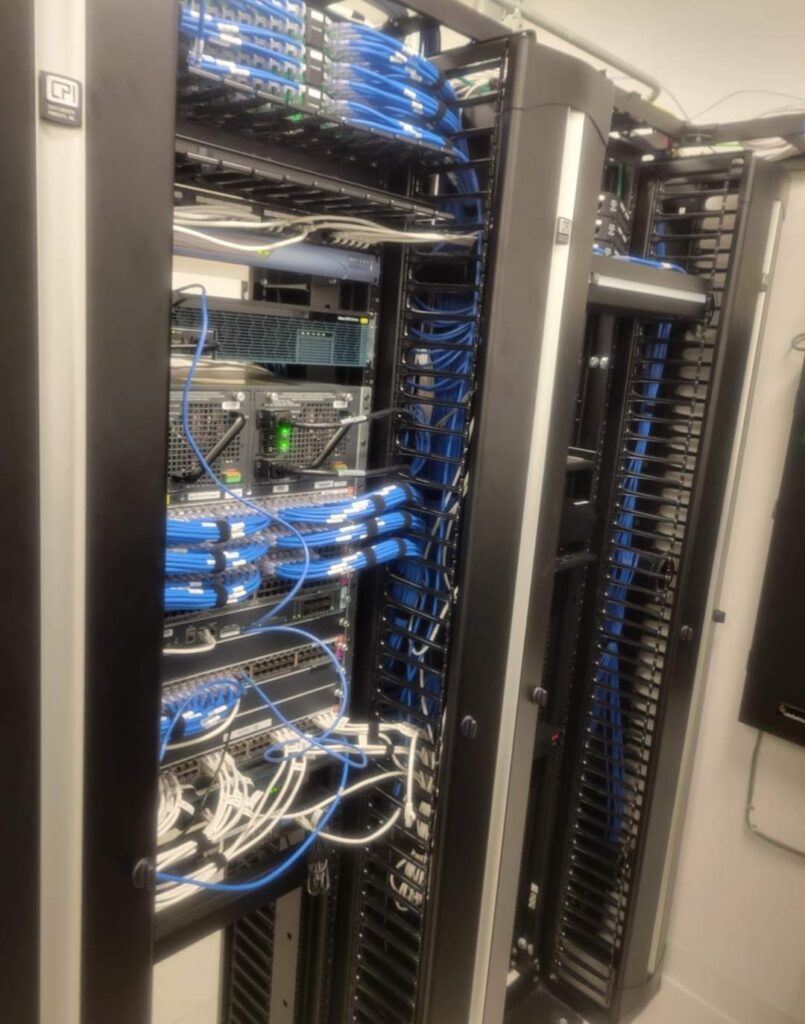 A server room with several racks filled with network equipment. Blue and white cables are neatly organized and connected to various devices. The room is well-lit and the equipment appears modern and maintained.